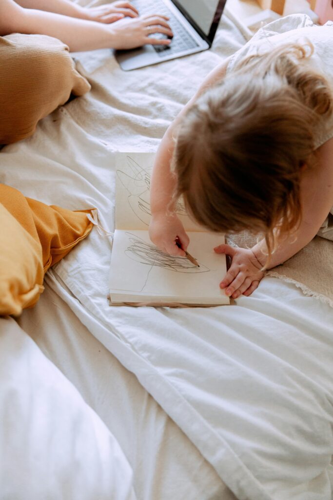 Girl drawing in notebook while mother working remotely nearby // Healthier Baby Today