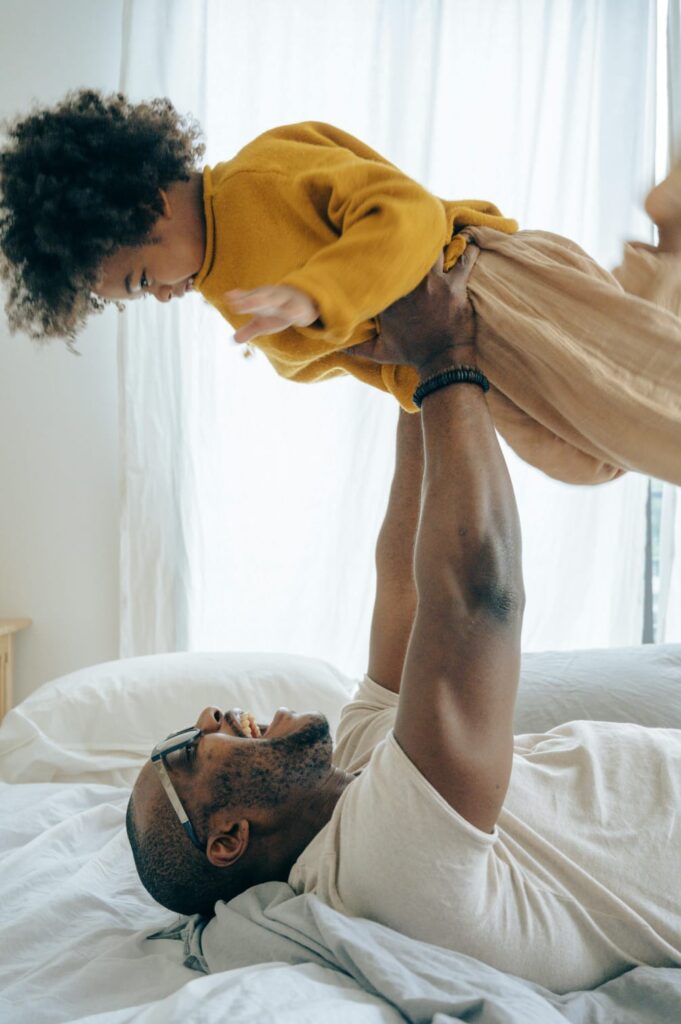 dad playing with toddler, wearing glasses, laying on bed, lifting girl // Healthier Baby Today