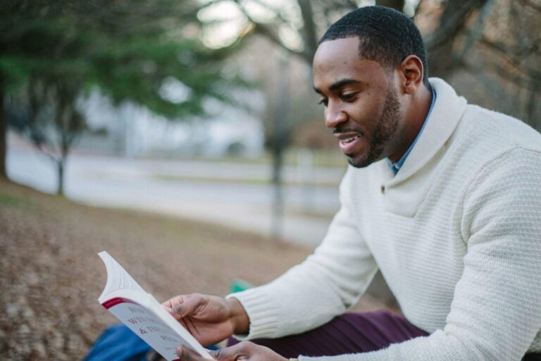 man reading book at park, white and red book, man smiling, baby book // Healthier Baby Today