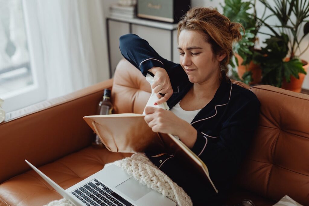 Woman sitting on couch in front of laptop, tearing a page out of a book, feeling frustrated // Healthier Baby Today