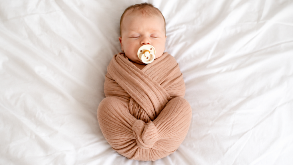 Sleeping newborn baby swaddled snugly in a brown blanket with their knees and legs curled toward their body, showing the tucked posture commonly known as the newborn scrunch // Healthier Baby Today